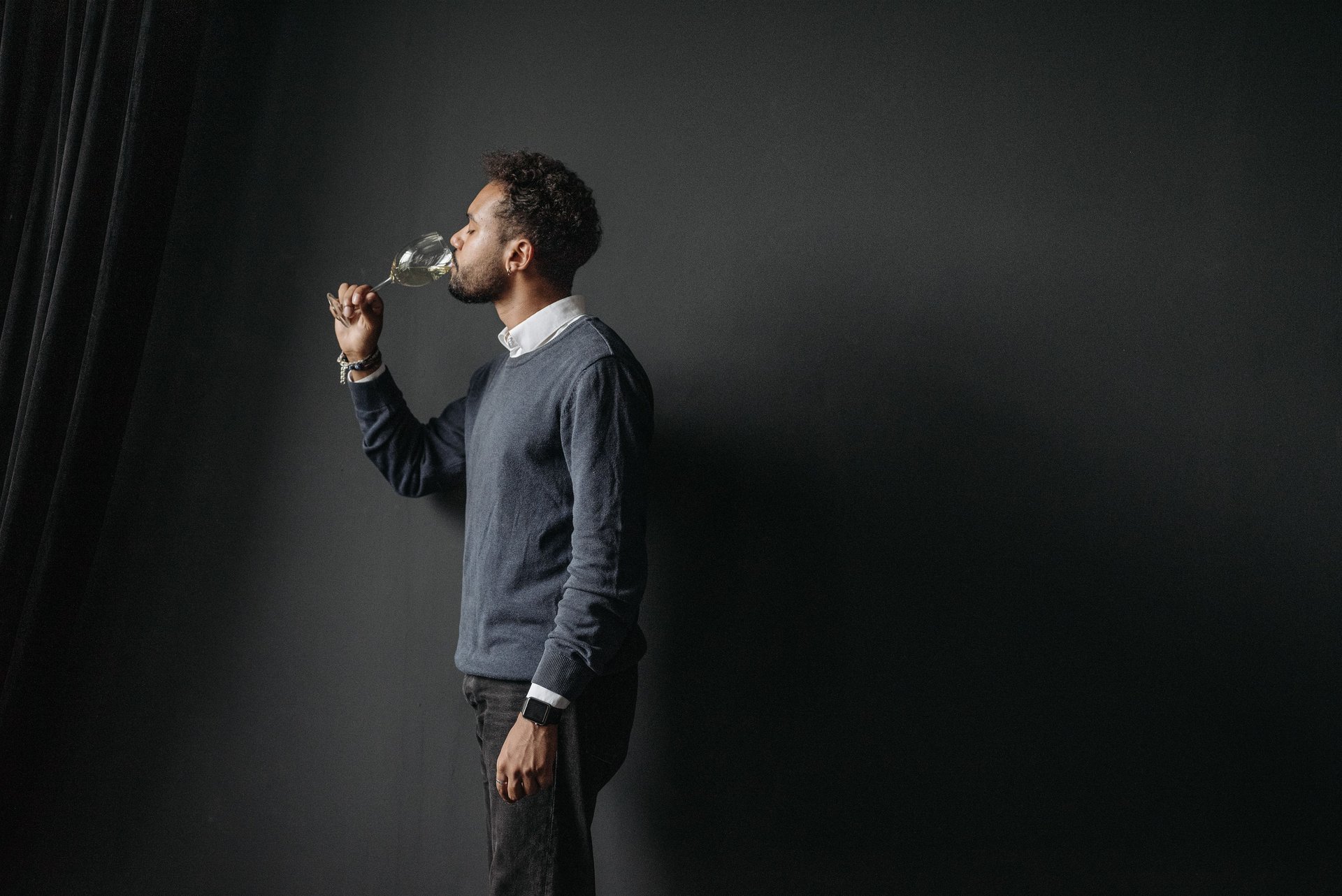 A man in a grey sweater closes his eyes and noses a glass of white wine against a dark background, checking it before the first sip