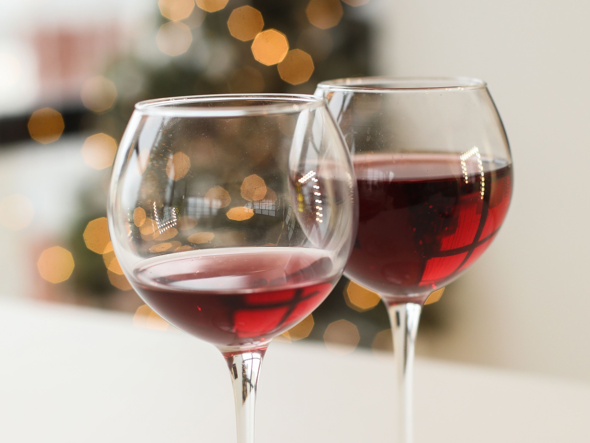 Two stemmed glasses of red wine standing side by side on a white table, with warm blurred lights in the background