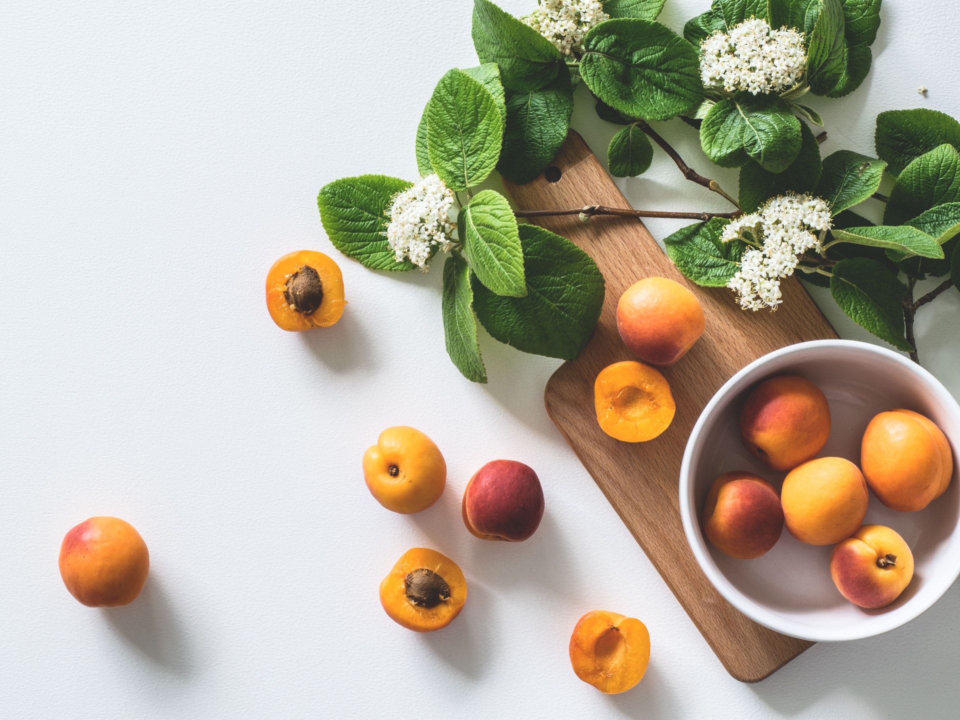 Fresh apricots, some of them halved, in a white bowl and on a wooden board, beside sprigs of white blossom and green leaves on a pale surface